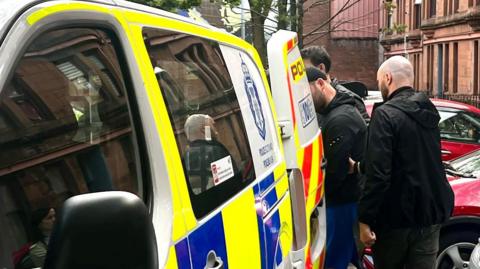 A man is escorted into the back of a blue, white and yellow Police Scotland van by two plain-clothes police officers on a residential street. Cars are parked nearby.