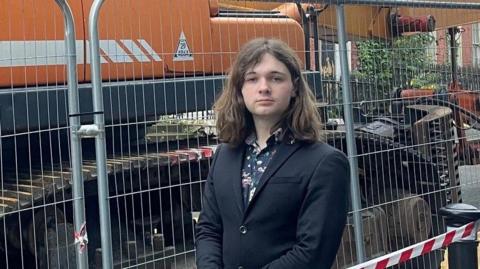 Ciaran Bell has long brown hair and is standing in a suit near a large crane. 
He is close to a barrier and a number of bollards with red and white tape near it. 