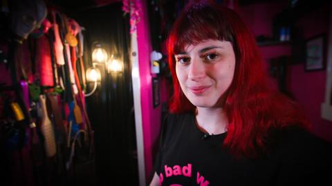 Porcelain Victoria is smiling at the camera. She has long hair, dyed bright red hair. She is wearing a black T-shirt with bright pink text on it. On the left hand side there is a collection of straps and paddles, arranged on hooks. The room is dark.