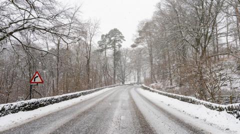 The A591 in Grasmere is a two lane rural road, with stone wall barriers on either side and a red sign indicating a sharp bend ahead. The road is dusted with snow, as are the tall trees bordering the road. Snow is falling.