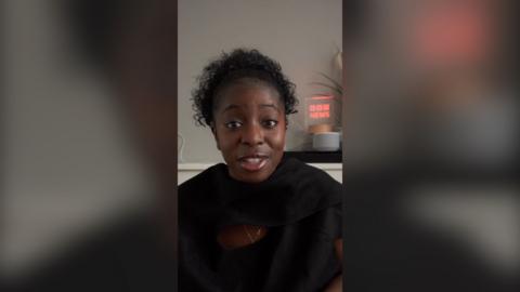 A woman with curly black hair wearing a black top talks to the camera
