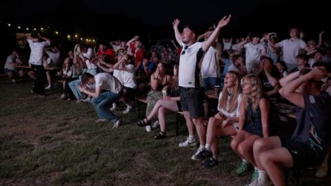 A large outdoor crowd gathers on long benches at night, many with hands on heads or arms raised, reacting intensely to a dramatic moment during an England v Ghana match at the 2026 World Cup.