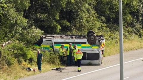 An NHS ambulance on its roof by the side of the road.