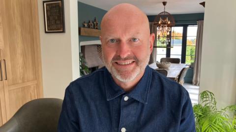 Kev Griffiths is smiling while sitting in the kitchen of his home. He is wearing a denim shirt. He is bald and has a grey beard.