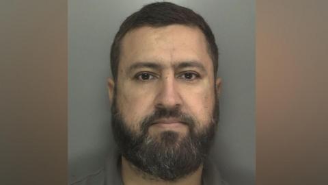 Shahid Adnan has dark hair and a beard  and wears an open-necked khaki shirt.