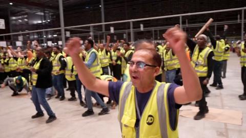 Warehouse workers wearing yellow vests celebrate with hands in the air