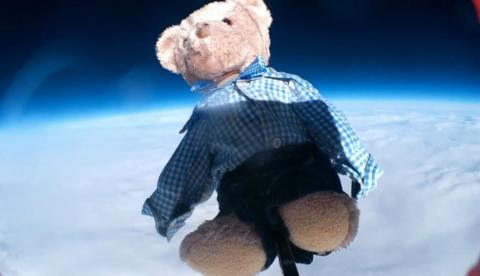 mall, brown bear, wearing school uniform, navy blue shorts and a blue and white checked shirt flying attached to a weather balloon above clouds