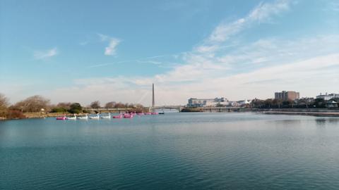 A wide expanse of water with pink and white flamingo boats to right, buildings and a bridge in distance under a blue sky 
