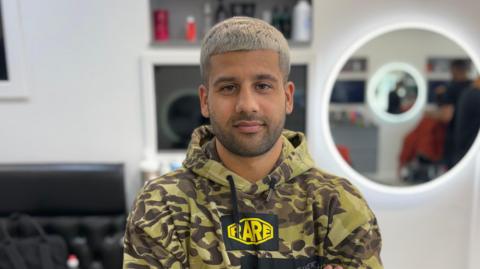 Ahmed wears a camouflage hoodie with a yellow logo. He has bleach blonde hair and faces the camera and is standing against the background of the salon