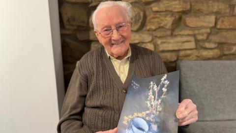 David Hughes, wearing thin-rimmed silver glasses, and with a brown cardigan on top of a yellow shirt, sits down. He is holding a copy of one of favourite artworks, which shows a selection of plants against a glass bowl. In the background is the beige brick from Washington Arts Centre.