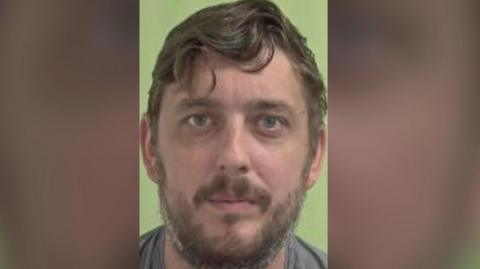 A mugshot of Liam Dunne, a man with wavy mouse brown hair and a beard, there is a green background.