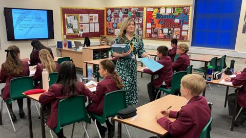 A teachers hands out blue workbooks to children in purple blazers sat at their desks in a classroom.