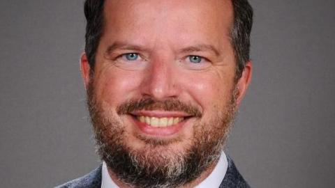 A studio photo of Anthony Griffin. He is smiling and has bright blue eyes and a grey and brown beard and moustache with grey and dark short hair.