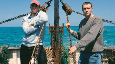 A scene from the film in which George MacKay and Callum Turner are standing on a fishing boat while at sea on a sunny day. MacKay is holding on to a chain fed into a pulley while Turner leans on a chain with his arms crossed. They are both looking steadily ahead. 