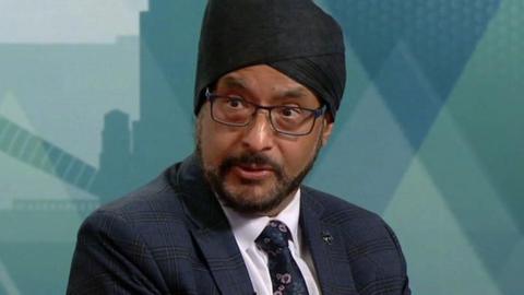 A man in a navy suit and black turban. He is sitting on a sofa and looking to the left. He is wearing a white shirt and floral tie, as well as rectangular glasses.
