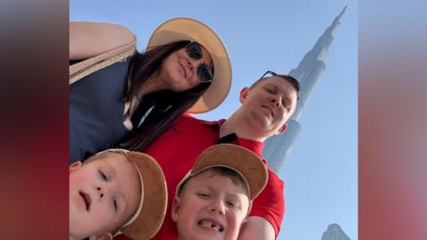 Pete Williams is smiling as he takes a selfie with his partner and two young boys in front of a skyscraper. He is wearing  red t-shirt and has short brown hair and  a pair of sunglasses over his head.  His partner has long straight dark hair and a beige brimmed hat. She is wearing sunglasses and and blue vest. The boys have short hair and are wearing light brown caps. 