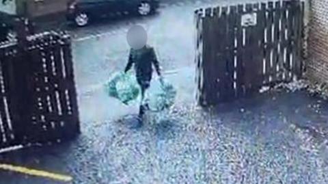 A man carrying four bags of rubbish, two in each hand. He is walking through an open gate into a driveway, with wooden fencing either side. His face is obscured. 