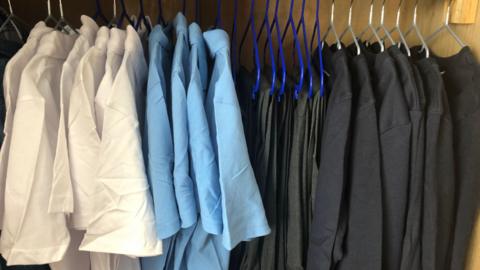 A row of neatly hung school uniform inside a wooden wardrobe. The shirts are arranged by colour, starting with white on the left, then light blue, then dark grey trousers, and finally black jumpers on the right. 