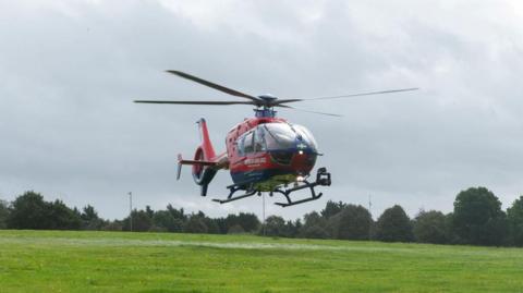 A red helicopter hovering above a green field on a cloudy day.