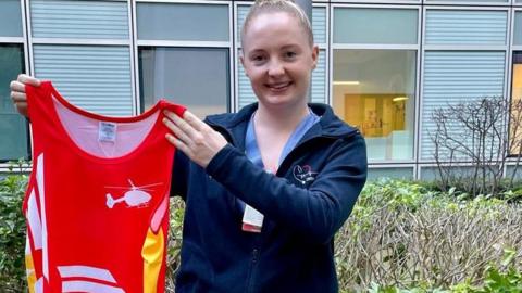 Emily wearing a blue fleece holding a red jersey, smiling for camera 