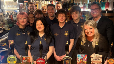 A number of staff are behind the bar in their uniform next to the owners.