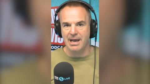 Martin Lewis wearing a green tshirt with a black 5 Live branded mic below him.