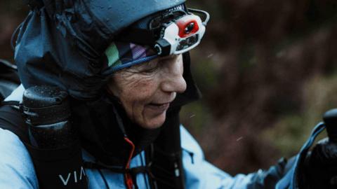 Runner Anna Troup during the Spine Race. There is a blue-ish wash on the photo, which depicts Anna mid-running in rainy conditions. She is wearing a blue waterproof and a navy headband and a headtorch.