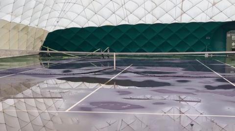 The inside of a white dome. There are tennis courts inside with nets. There are large puddles around the dome. 