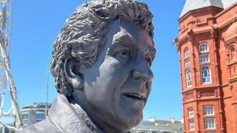Statue of Rhodri morgan is grey metal, his face looking upward slightly with his mouth open as though he is about to speak and you can also see the collar of an overcoat. the red pierhead building and the ferris wheel attraction at mermaid key are in the background on a blue sky day