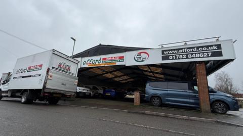 A garage with vehicles parked under a large roof. A white van is parked at the side of the road in front of it.