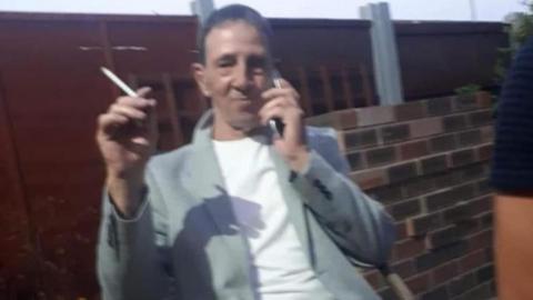David Aubert, in his 50s, who has dark blonde hair, sits on a chair in a back yard holding a cigarette in his right hand and a mobile phone to his ear in his left hand.
He is wearing a light grey blaze over a white t-shirt.
