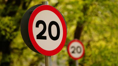 A 20mph sign in the fore with another out of focus behind it, with a backdrop of green trees.