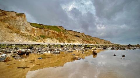 Side of cliffs from sandy shore with stones in wet sand. Cloudy, grey sky