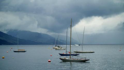 Boats out on an inlet with blue grey sea, mountains behind and huge grey rain clouds in the sky