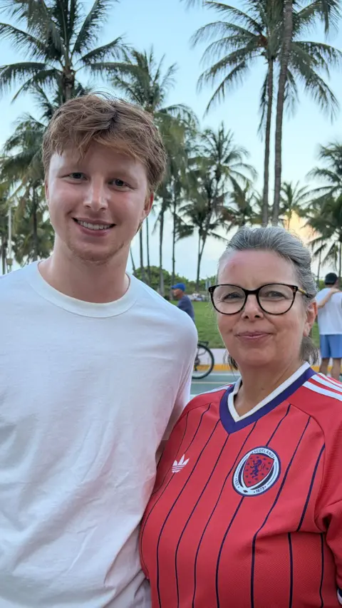 A young man and a slightly older woman stand side by side outdoors, smiling at the camera. The man has short light‑brown hair and wears a plain white T‑shirt. The woman has grey hair tied back, wears glasses, and is dressed in a red Scotland football shirt with thin blue stripes. Behind them are tall palm trees and people walking or cycling along a path in warm evening light.