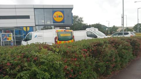 A photo of a large Lidl supermarket. Police vehicles are parked in the car park, including one marked "forensics". A police cordon is visible. A hedge surrounding the car park sits in the foreground. 