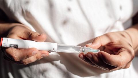 A close up of a health professional, in white clothing. They're holding a weight loss drug - in the form of a tube or syringe - that is ready to be injected.