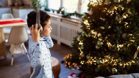 A young boy wearing blue pyjamas standing to the left of the photo. He is reaching up and touching his headphones while looking at the Christmas tree in front of him, which is lit up with gold lights and green baubles. Behind him is a dining table and a green garland resting on the windowsill. 