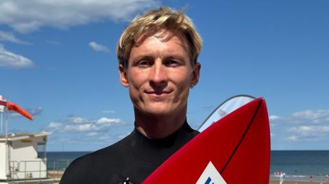 Louis Hudson is standing in front of the sea and an orange RNLI flag can been seen to the right. He has blonde tousled hair and is holding a red surf board with a black stripe dow the middle. He has a tanned complexion, has a hoop gold earring and is wearing a black wetsuit. The cloudy, blue sky can be seen in the background as well as the slightly out of focus water.