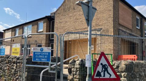 View of a boarded up two-storey building, There is a metal fence surrounding the site and a "men at work" road sign.