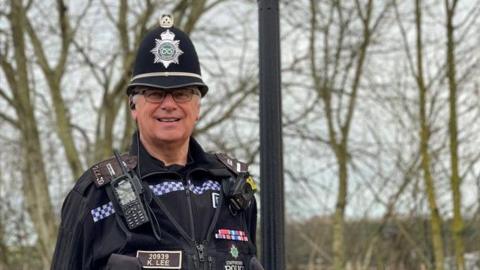 Police officer Kevin Lee is stood next to a lamppost. He is wearing glasses and black police uniform with a body-worn camera, police radio and other police equipment attached. There are trees in the background.