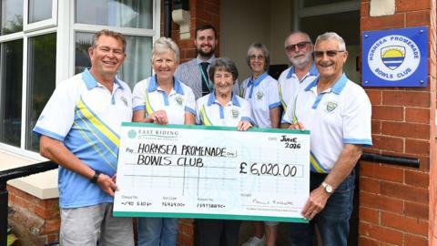 A group of three women and four men stood outside Hornsea Promenade Bowls Club. Three women and three men are wearing blue, white and yellow bowls kit. The other man is wearing a striped, blue shirt and a tie. Some of them are holding a large check for £6,020.00 written to Hornsea Promenade Bowls Club.
