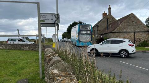 A rural crossroads with a car pulling in front of a bus while another car is waiting to pull out. Behind the bus is a long line of traffic.