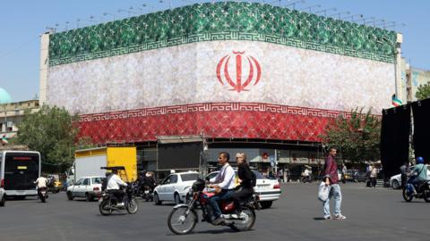 Iranians drive cars and motorbikes past a billboard featuring Iran's national flag at Enqelab Square in Tehran, Iran.