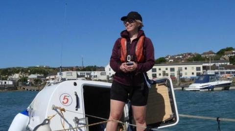 Nicky Allen. wearing a burgundy jacket, black rowing shorts a black cap and dark glasses, stands on a boat in the sea. She is smiling but not looking at the camera.