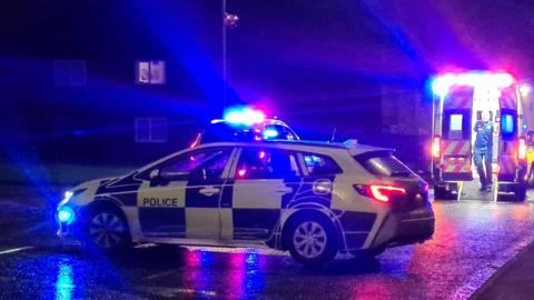 A blue and yellow checked police car with its sirens on parked across a dark road. There is another emergency vehicle behind it with its lights visible. To the right is an ambulance with a man walking down the ramp. There are houses on the left.