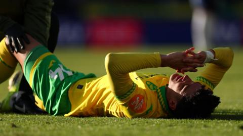 A photo of Ben Chrisene lying on the grass injured wearing a Norwich City home kit and a bandage around his wrist