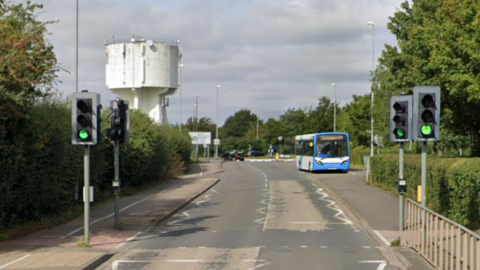 Street view of the road heading towards the roundabout on a cloudy day. There is a set of traffic lights and a crossing with bushes at either side of the road. In the distance is a white road sign for the roundabout.