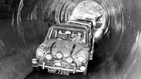 Minis in Coventry sewers during the filming of The Italian Job film.  A car with five headlights drives in a black and white photo in a concrete pipe with other cars behind it.