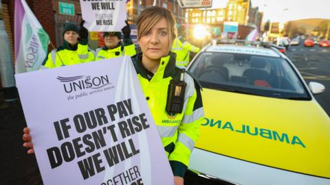 An ambulance worker in florescent jacket holds a sign saying "if our pay doesn't rise we will" standing beside an ambulance estate car. Behind her are other striking ambulance workers. 